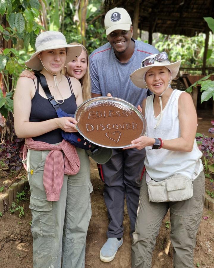 Four people smiling and holding a tray of ground coffee with a message written in it, under banana leaves