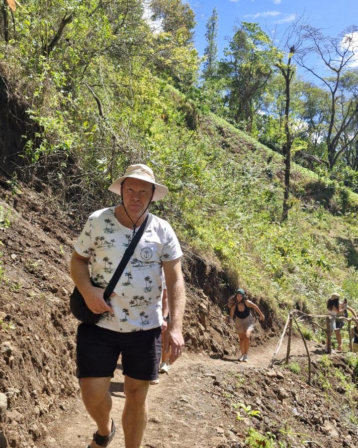 A hiker in a sun hat walking up a dirt trail through green forest on a steep hillside